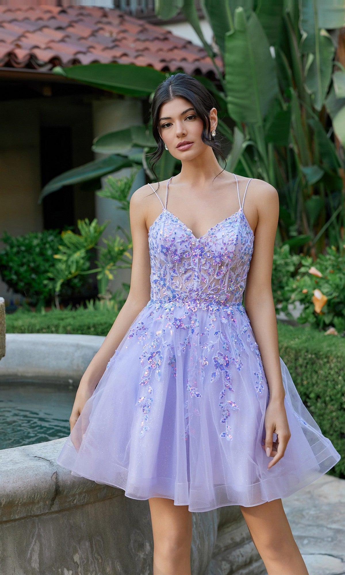A woman stands outdoors by a fountain wearing the Juliet JT988N short homecoming dress, featuring a lilac fit-and-flare silhouette with thin straps and floral embroidery. Lush green plants are in the background.